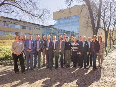 Zu sehen sind eine Gruppe von Menschen neben Bäumen und vor einem Gebäude auf einer Wiese