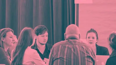 Auf dem Bild sind Personen zu sehen, die sich miteinander um einen Tisch herum unterhalten. Das Bild ist rötlich eingefärbt