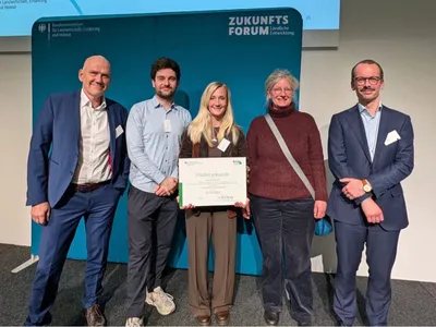 Fünf Personen stehen vor einer blauen Wand mit dem Schriftzug »ZUKUNFTS FORUM Ländliche Entwicklung«, eine Person hält eine Förderurkunde in der Hand.