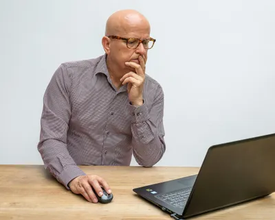 Ein Mann sitzt am Schreibtisch und schaut skeptisch auf seinen Laptop